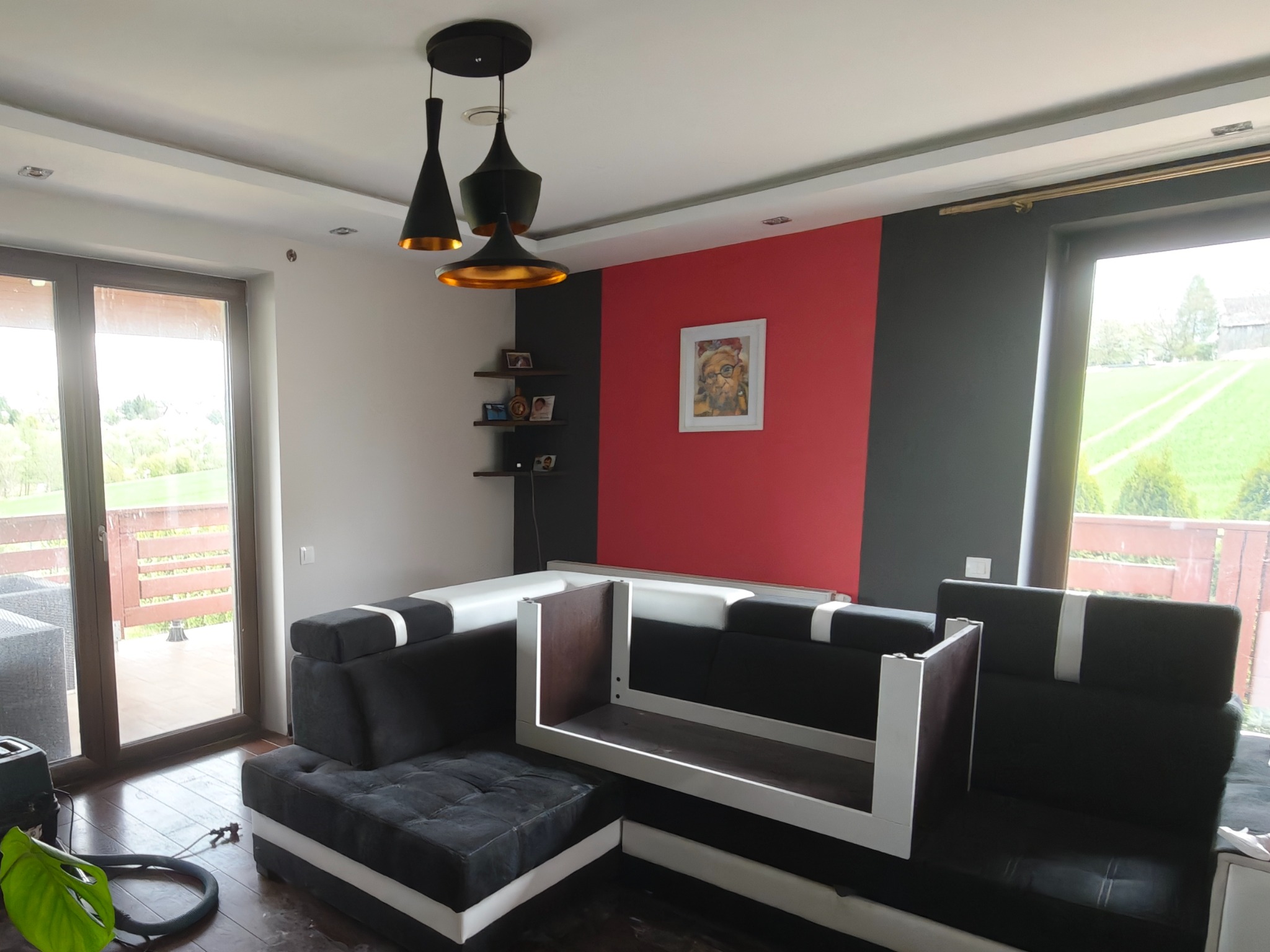 Salon po malowaniu: czerwona ściana za sofą, szara obok okna. Nowoczesna lampa wisząca. Widok na zieleń za oknem. Parkiet po czyszczeniu.