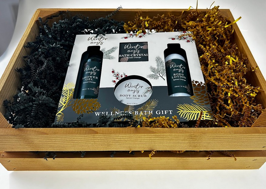 Drewniana skrzynka wypełniona czarnym i złotym papierem dekoracyjnym, w której umieszczony jest zestaw kosmetyków 'Winter Oasis Spiced Orange' zawierający żel pod prysznic, kryształki do kąpieli...