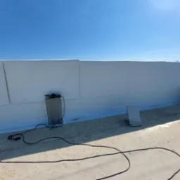 Białe płyty izolacyjne na dachu budynku w słoneczny dzień. Widoczne kable i narzędzia, betonowe elementy konstrukcyjne. Prace dekarskie w toku.