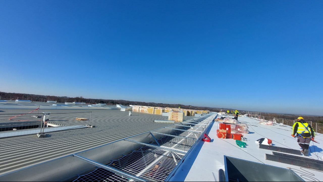 Prace na dachu hali: ekipa w żółtych kamizelkach montuje izolację i zabezpieczenia. Widok z góry, błękitne niebo.