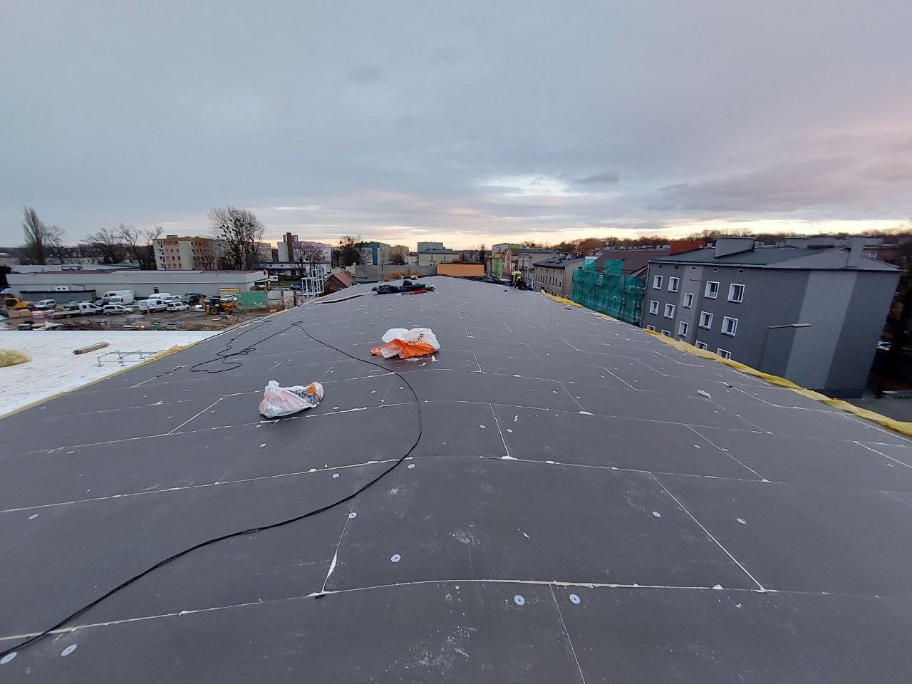 Płaski dach pokryty czarną membraną z widocznymi łączeniami, w tle zabudowa miejska i pochmurne niebo. Prace dekarskie, hydroizolacja dachu.