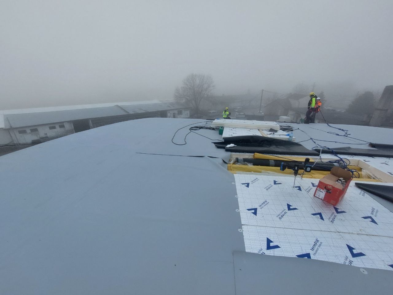 Montaż izolacji na dachu budynku w mglisty dzień. Widoczne narzędzia, materiały izolacyjne i pracownicy w odblaskowych ubraniach. Dach pokryty szarą membraną.