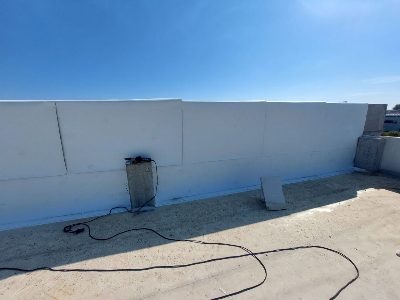 Białe płyty izolacyjne na dachu budynku w słoneczny dzień. Widoczne kable i narzędzia, betonowe elementy konstrukcyjne. Prace dekarskie w toku.