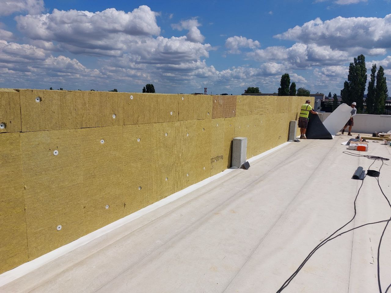 Montaż izolacji termicznej na dachu płaskim z widocznymi mocowaniami i ekipą pracowniczą układającą kolejne płyty. Błękitne niebo z chmurami w tle.