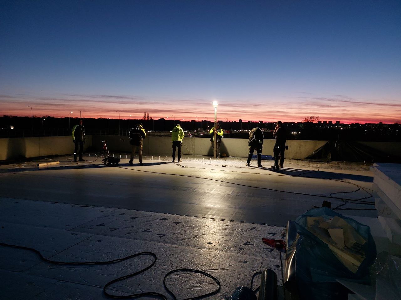 Prace dekarskie na dachu o zmierzchu: ekipa w odblaskowych ubraniach, rozłożone materiały izolacyjne, widok na miasto w tle pod rozgwieżdżonym niebem.