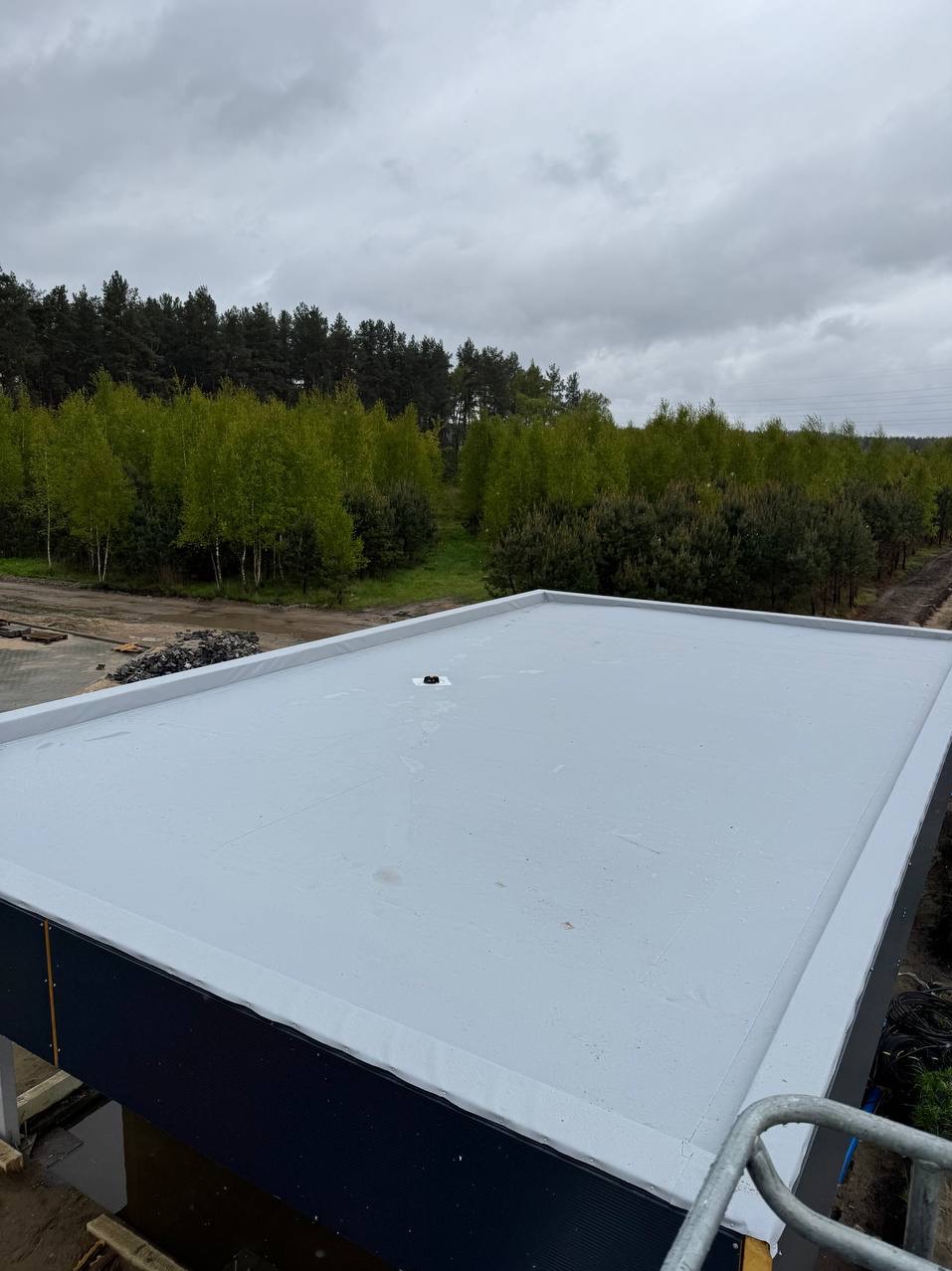 Szary, płaski dach budynku z widocznym odpływem, na tle lasu i pochmurnego nieba. Ujęcie z góry, ukazujące gładką powierzchnię i detale wykończenia.