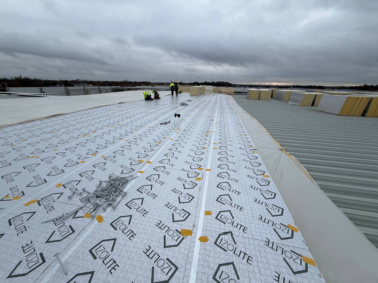 Montaż izolacji termicznej Izolite na dachu przemysłowym. Widoczni pracownicy w odblaskowych kamizelkach i narzędzia. Pochmurne niebo w tle.