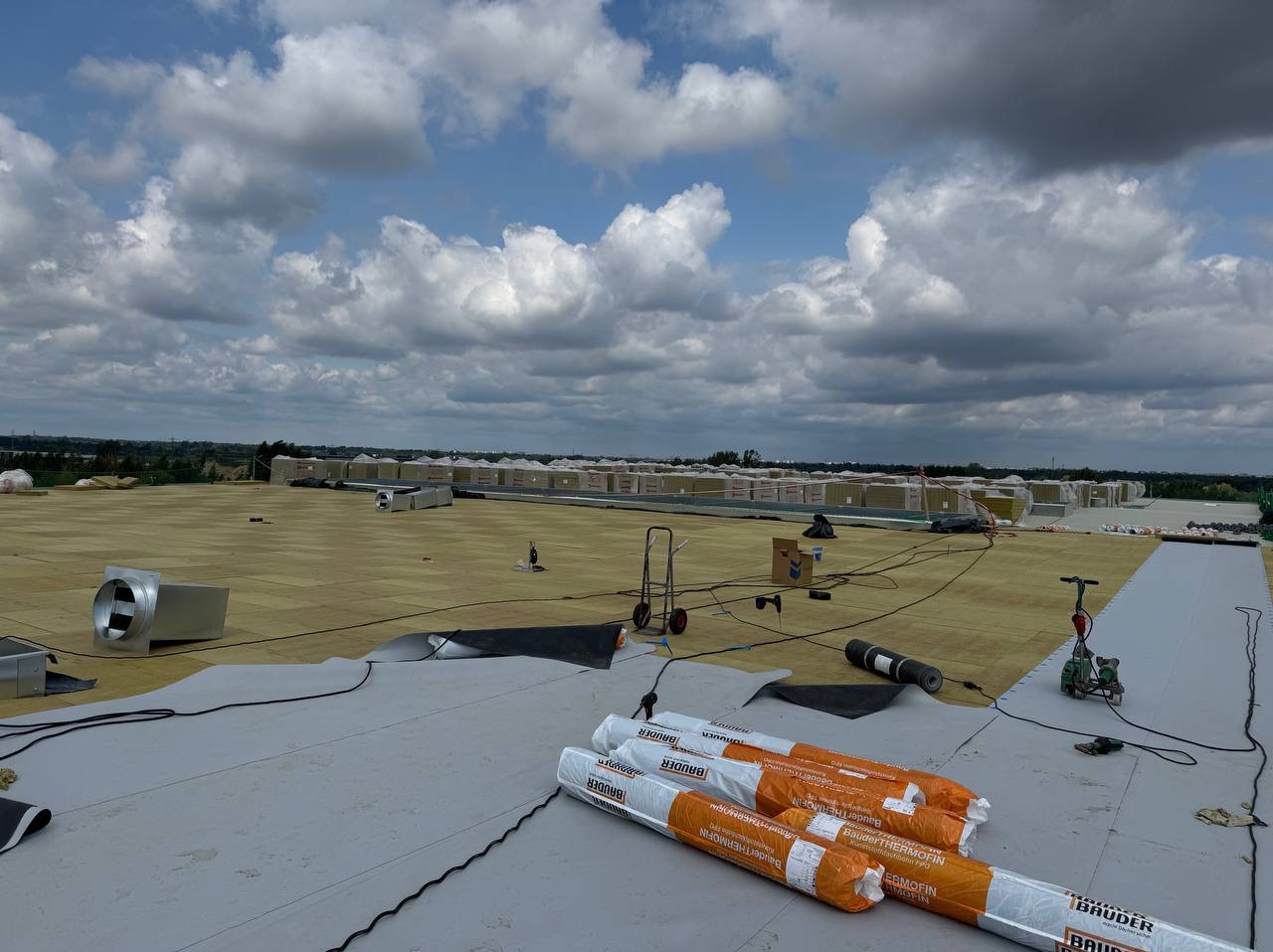 Rozległy dach płaski w trakcie izolacji termicznej, widoczne rolki materiału izolacyjnego Bauder Thermofin, wentylacja i narzędzia. W tle zapasy materiałów pod błękitnym niebem.