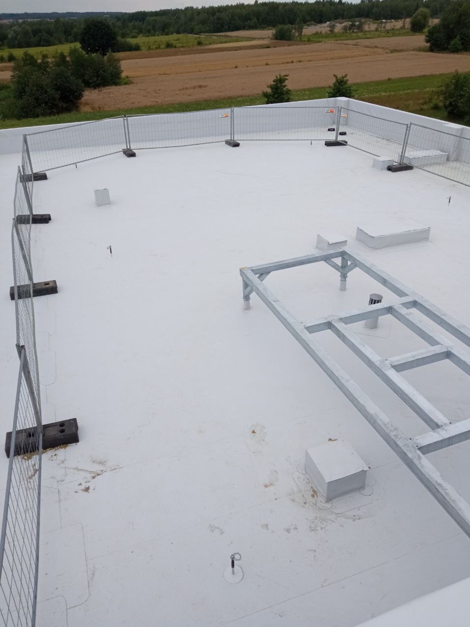 Biały dach płaski z metalową konstrukcją i zabezpieczeniami. Widok z góry na nową izolację, elementy wentylacyjne i krajobraz w tle.