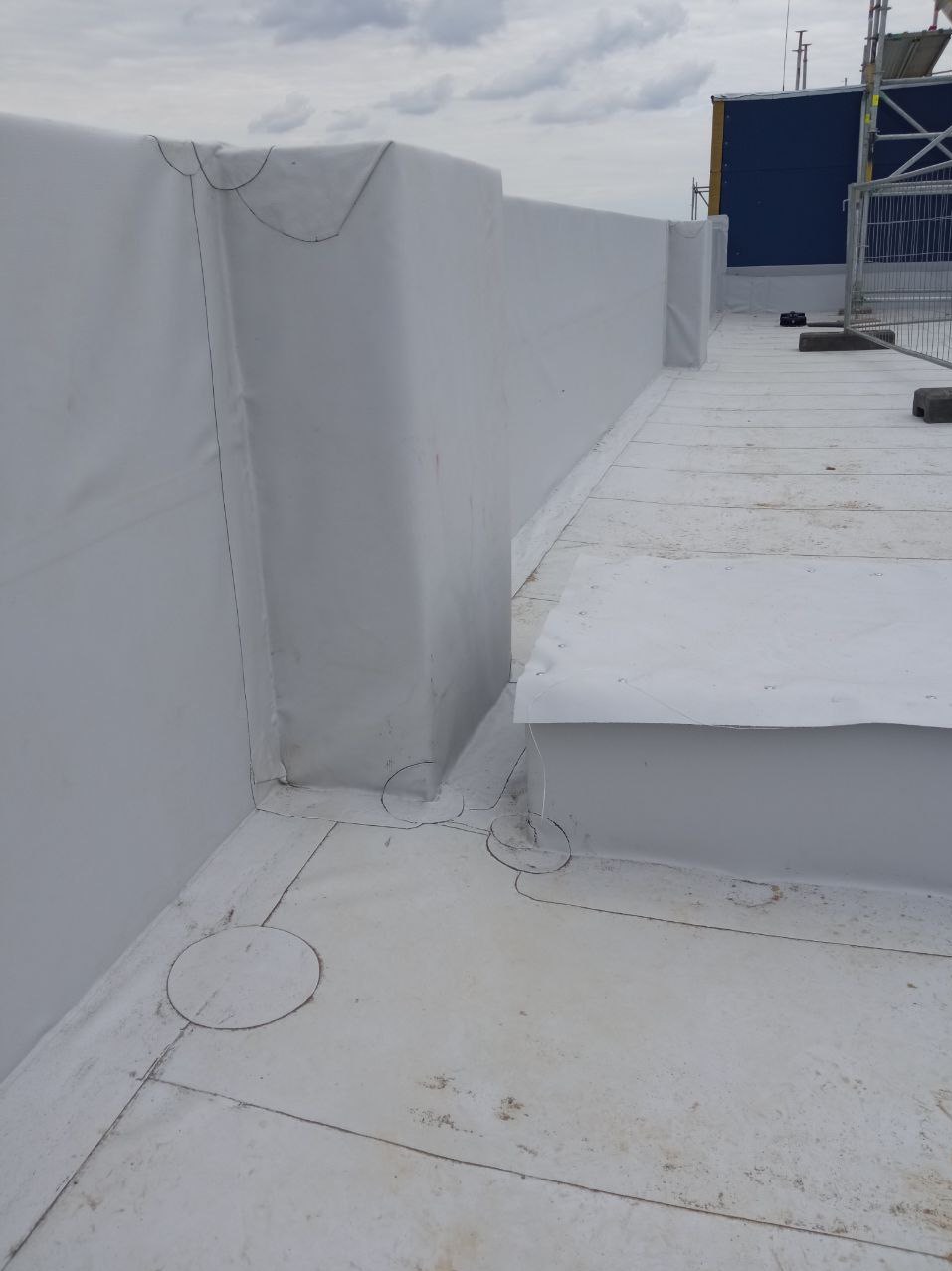 Biała membrana dachowa szczelnie pokrywająca płaski dach z wystającymi elementami, zabezpieczona na krawędziach. Widoczne łączenia i detale wykończenia.