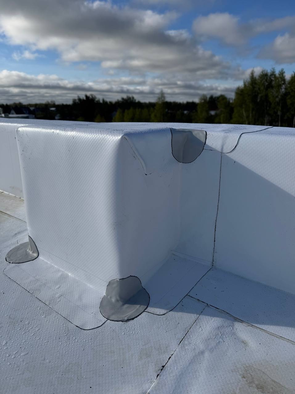 Szczegółowe ujęcie białej membrany dachowej z uszczelnieniami na płaskim dachu, z widocznym tłem drzew i nieba. Ukazane detale hydroizolacji.