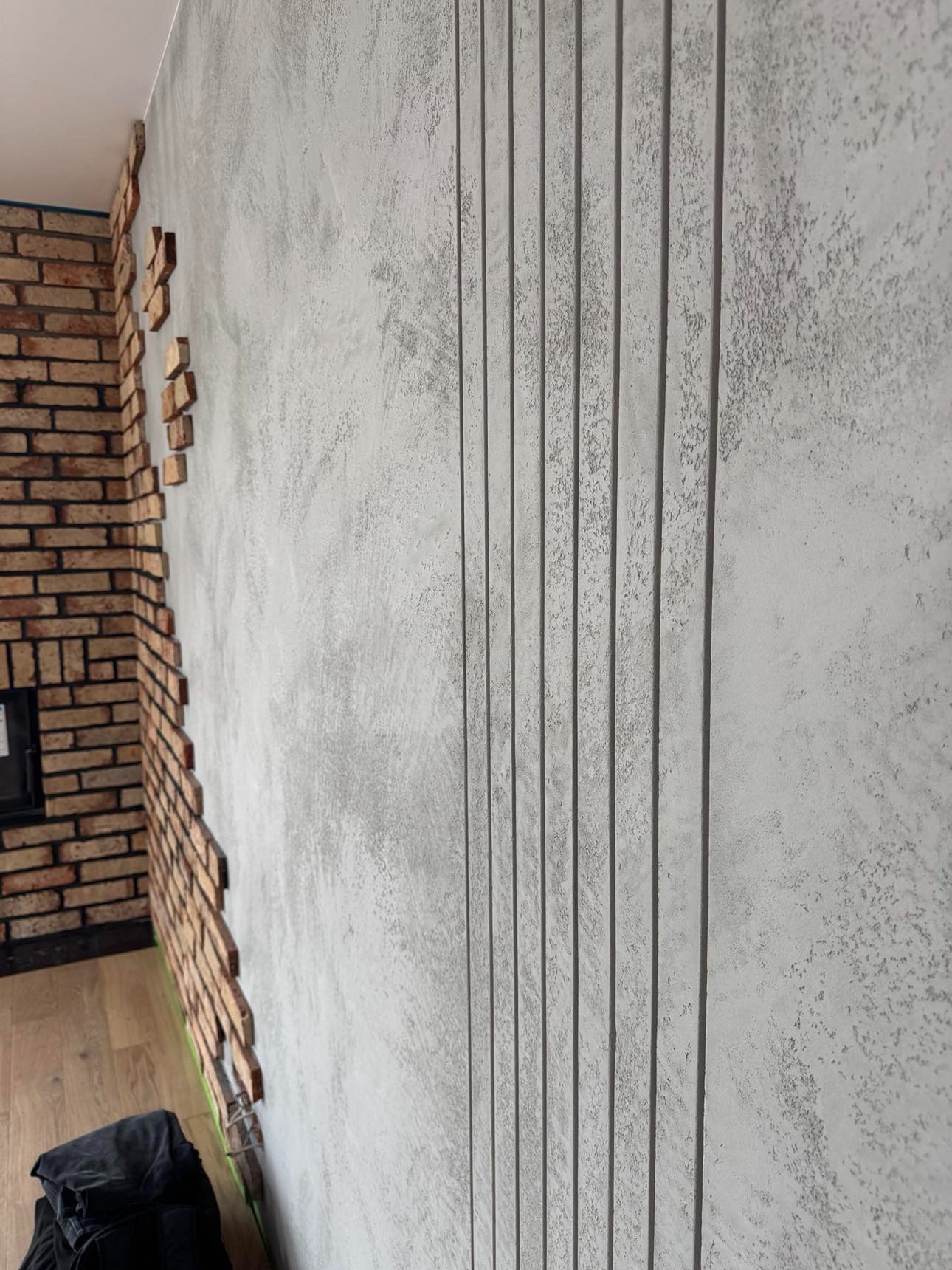 Ściana wykończona tynkiem dekoracyjnym z pionowymi wgłębieniami, obok fragmentu ściany w trakcie układania cegieł dekoracyjnych.