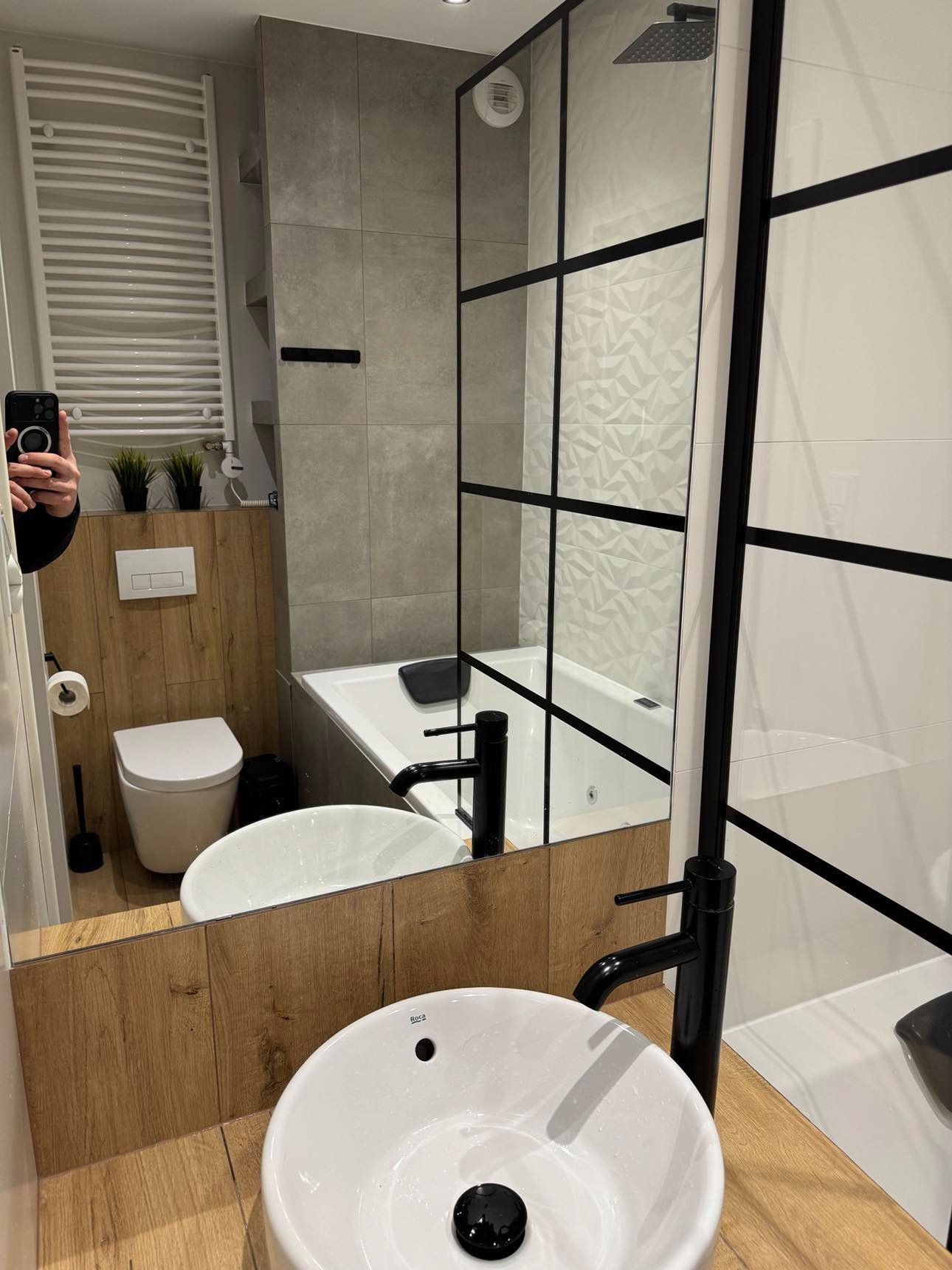 Nowoczesna łazienka z białymi sanitariatami Roca, kabiną prysznicową typu walk-in z czarnymi profilami i drewnopodobnymi panelami na ścianach.