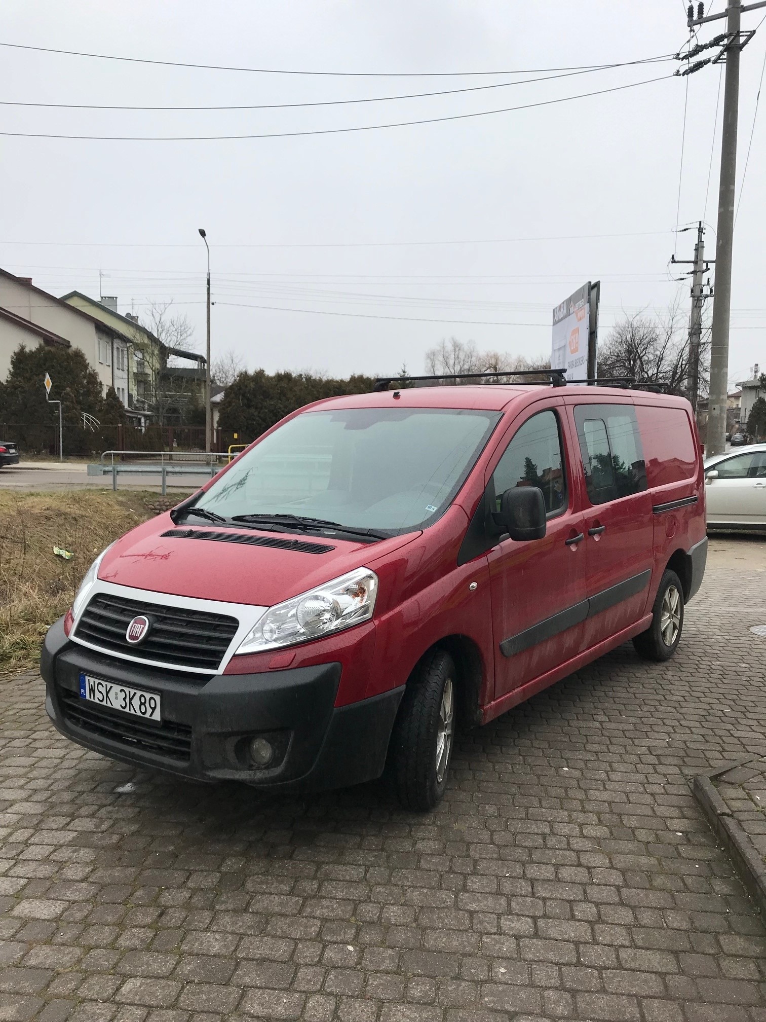 Czerwony bus Fiat Scudo zaparkowany na brukowanej ulicy w pochmurny dzień, z widoczną tablicą rejestracyjną WSK 3K89 i relingami dachowymi.