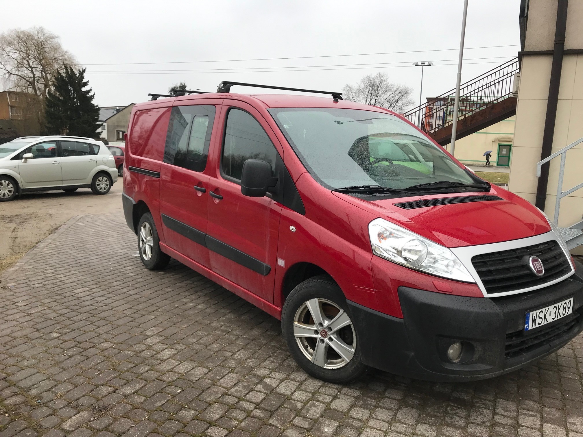 Czerwony bus marki Fiat Scudo z relingami dachowymi, zaparkowany na brukowanej nawierzchni, widoczny w pochmurny dzień.