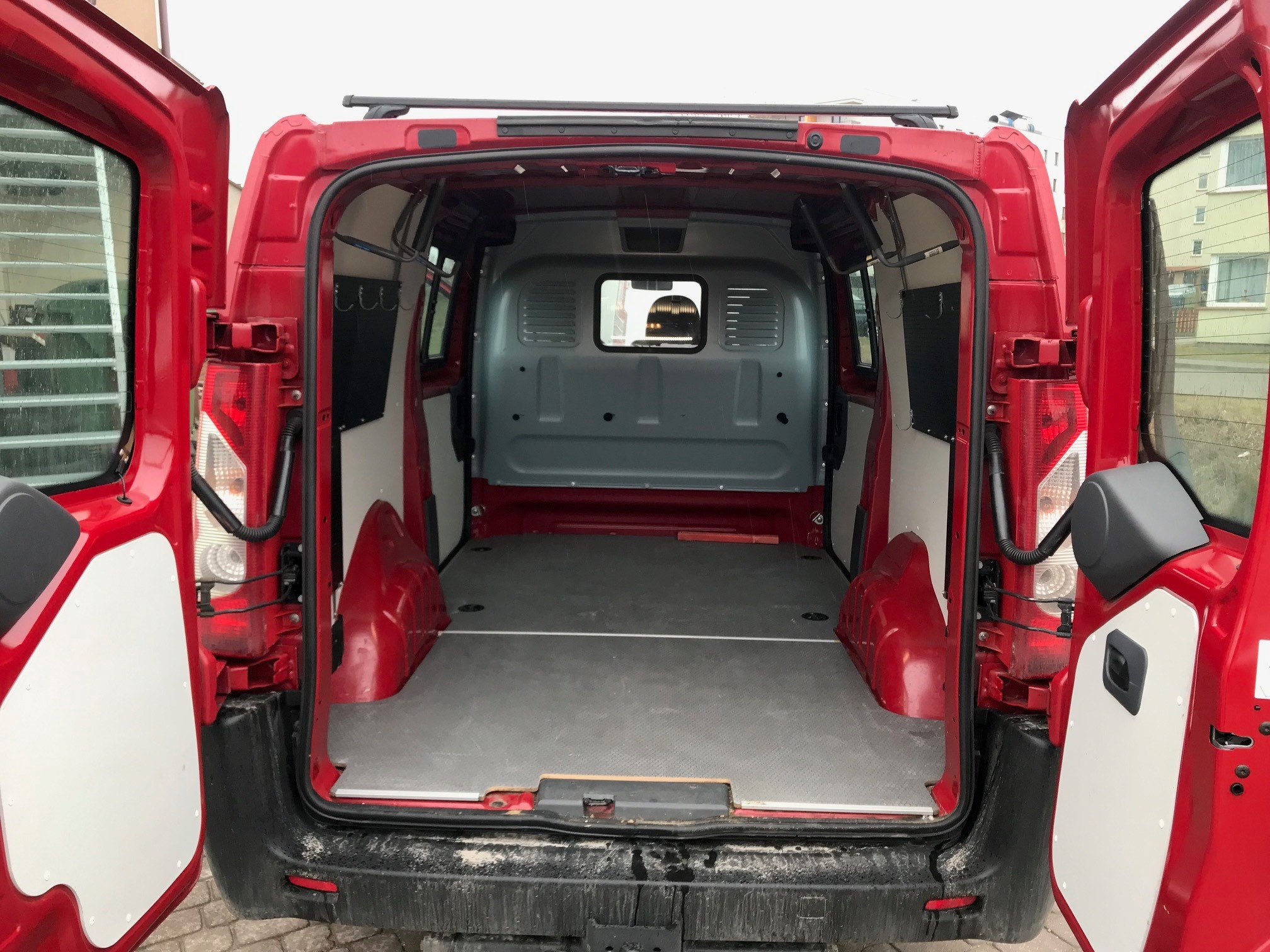 Wnętrze pustego, czerwonego busa z otwartymi tylnymi drzwiami, widok z zewnątrz na szarą podłogę i metalowe ściany z małym oknem z tyłu kabiny.