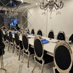 Elegancka jadalnia z długim stołem z białym blatem, krzesłami z czarną tapicerką i srebrnymi nogami, lustrzaną ścianą z geometrycznym wzorem i kryształowym żyrandolem.