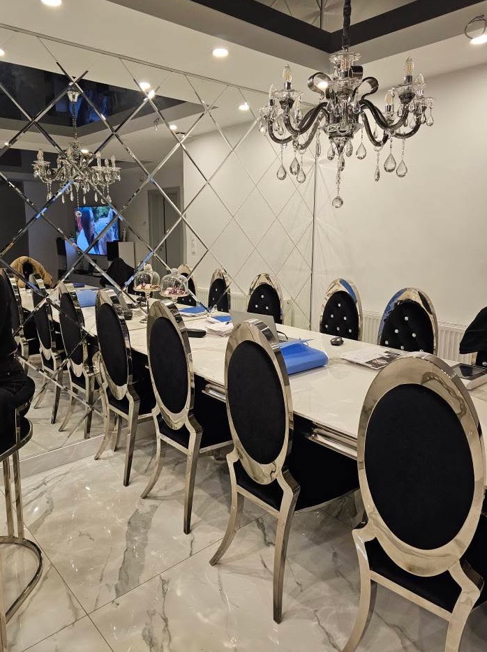 Elegancka jadalnia z długim stołem z białym blatem, krzesłami z czarną tapicerką i srebrnymi nogami, lustrzaną ścianą z geometrycznym wzorem i kryształowym żyrandolem.