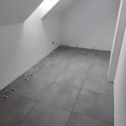 Szare płytki podłogowe w trakcie układania w pomieszczeniu ze skośnym sufitem i oknem dachowym, widoczne niebieskie krzyżyki dystansowe.