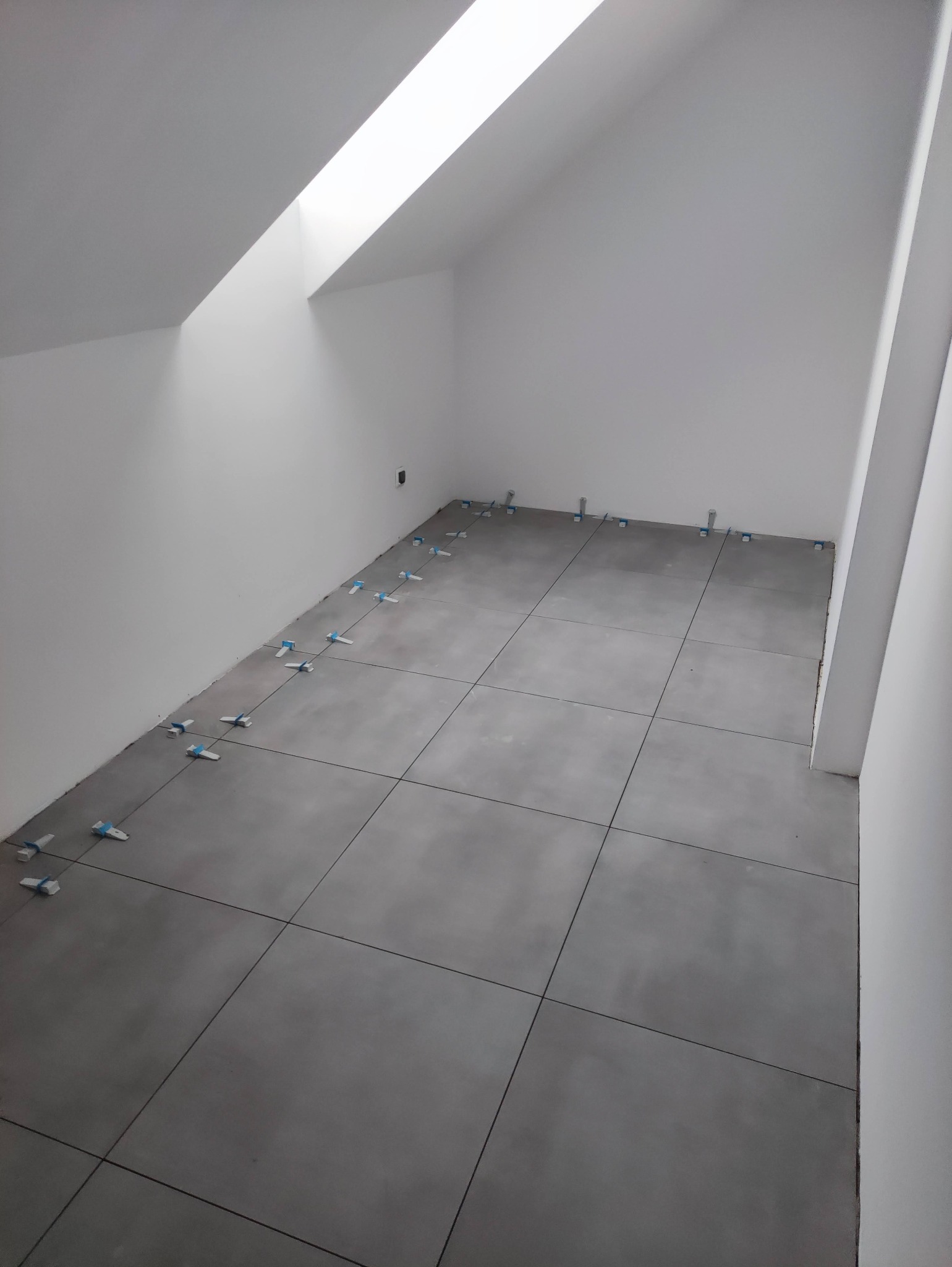 Szare płytki podłogowe w trakcie układania w pomieszczeniu ze skośnym sufitem i oknem dachowym, widoczne niebieskie krzyżyki dystansowe.