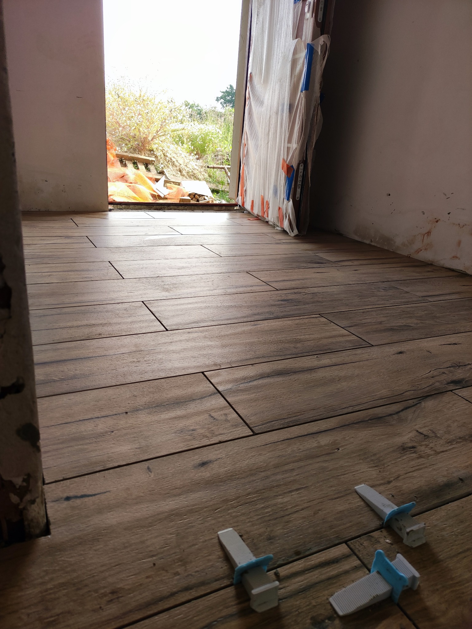 Ułożone podłużne płytki podłogowe imitujące drewno, z widocznymi plastikowymi klinami dystansowymi, w tle zabezpieczone folią drzwi balkonowe i widok na ogród.