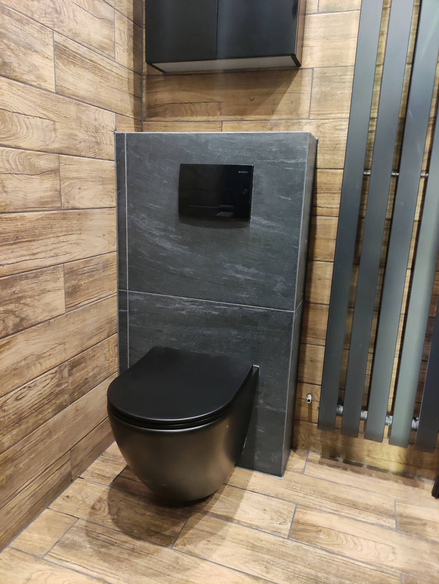 Nowoczesna, czarna, podwieszana toaleta z przyciskiem spłukującym Geberit, obudowana szarą płytą imitującą kamień, na tle drewnopodobnych płytek ściennych i podłogowych oraz grafitowego grzejnika...