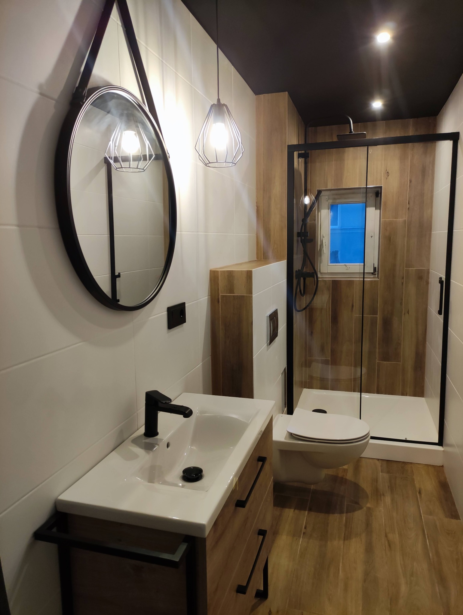 Nowoczesna łazienka z białymi płytkami, drewnopodobną podłogą i czarnymi akcentami: okrągłe lustro na pasku, minimalistyczna lampa, kabina prysznicowa typu walk-in z czarną ramą.