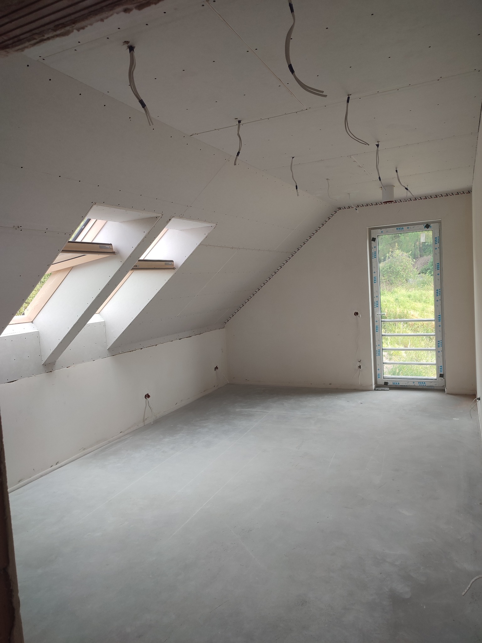 Poddasze w trakcie prac wykończeniowych: zamontowane płyty na skosach i suficie, widoczne okna dachowe, przewody elektryczne wystające z sufitu, ściany przygotowane do malowania, surowa wylewka...