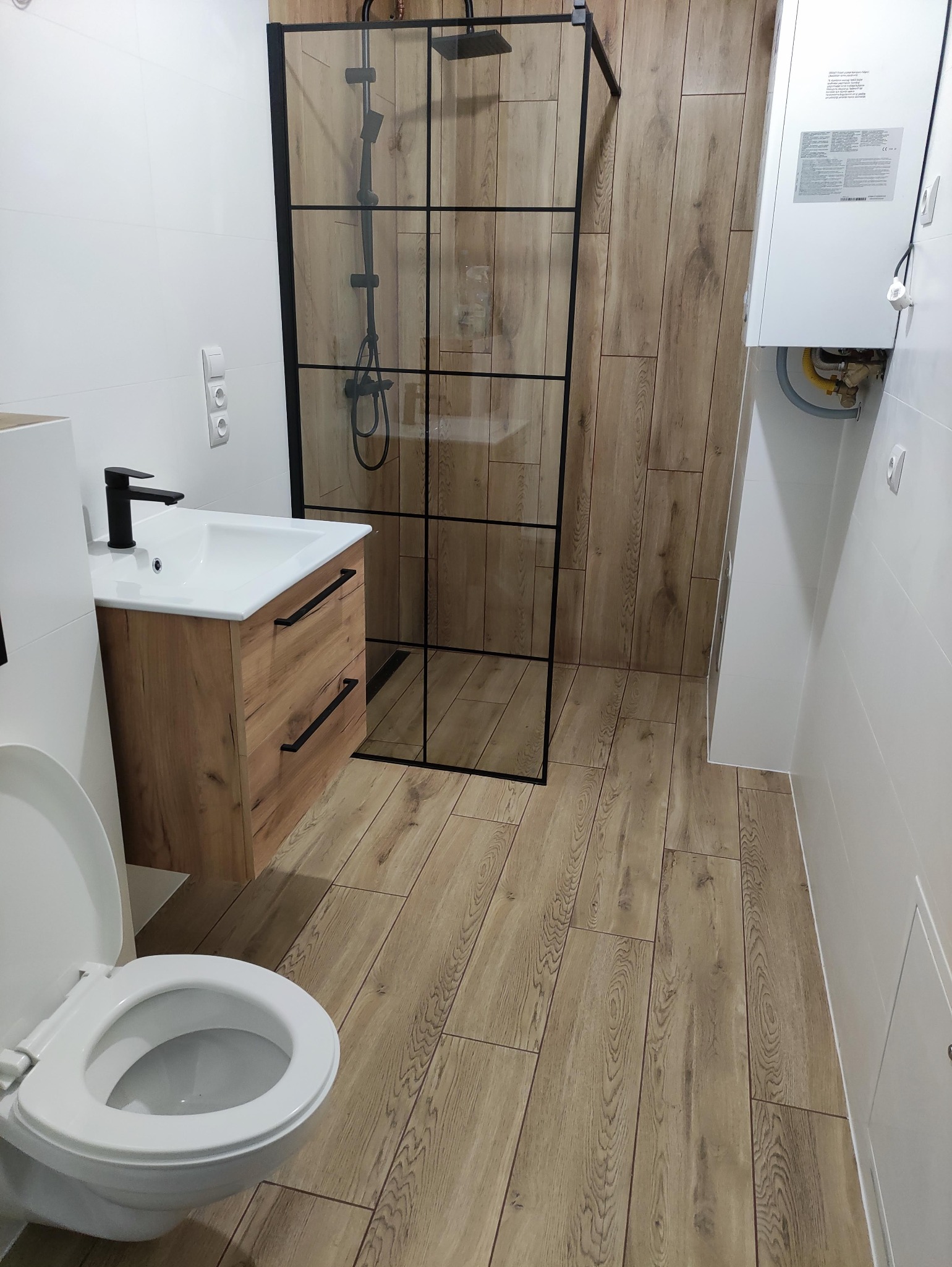 Nowoczesna łazienka z kabiną prysznicową typu walk-in z czarnymi profilami, umywalką z szafką w odcieniach drewna, białymi płytkami ściennymi i podłogą imitującą drewno, oraz wiszącym bojlerem.