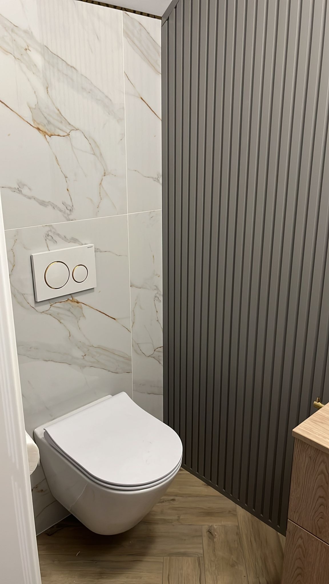 Nowoczesna toaleta podwieszana z białym przyciskiem spłukującym z elementami w kolorze złotym na tle marmurowych płytek oraz dekoracyjnej ściany z pionowych paneli w odcieniach szarości...