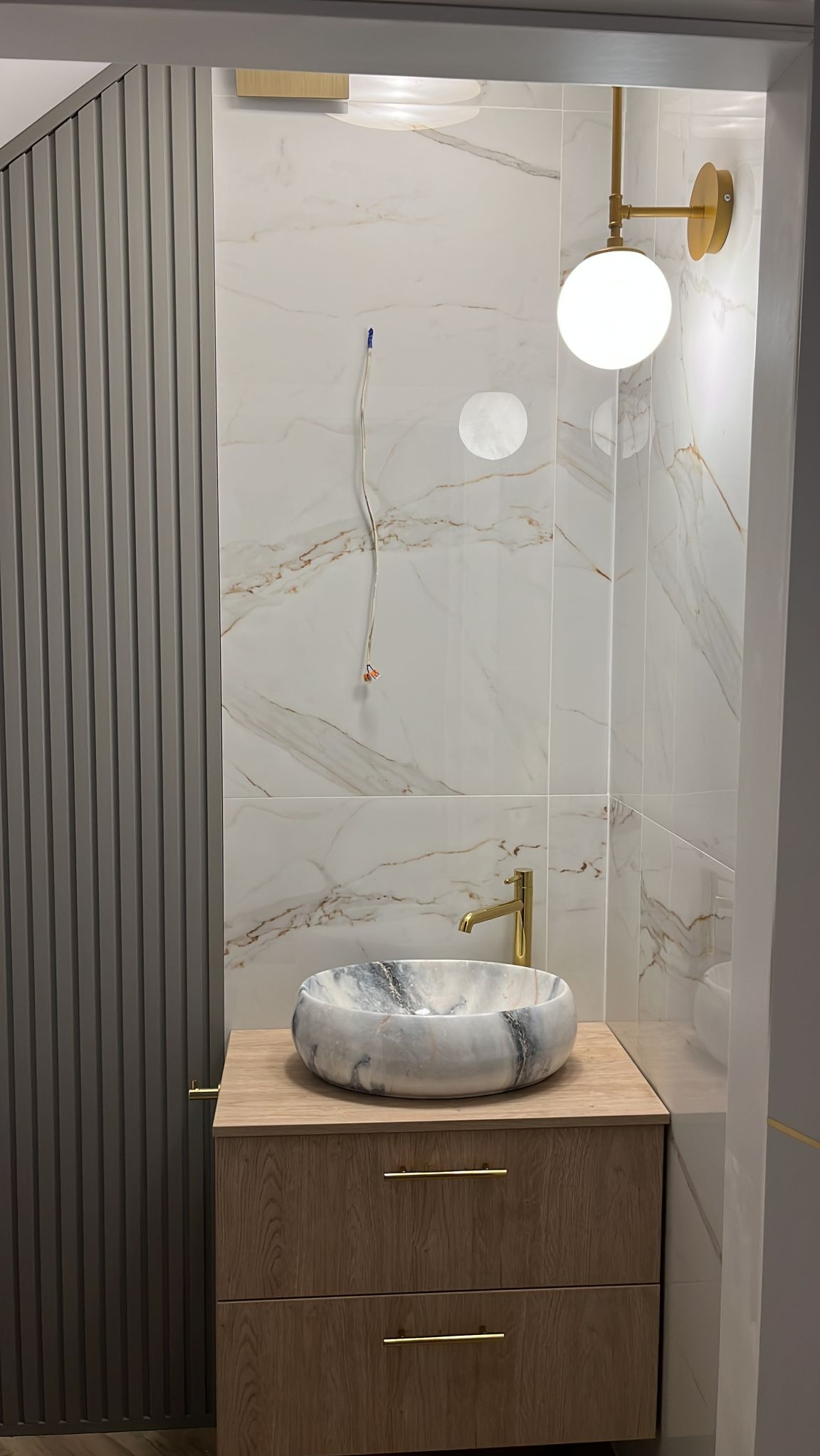 Nowoczesna łazienka z marmurową umywalką nablatową, złotą armaturą i oświetleniem, płytki imitujące marmur na ścianach, dekoracyjne panele ścienne w kolorze szarym, widoczne kable elektryczne przed...
