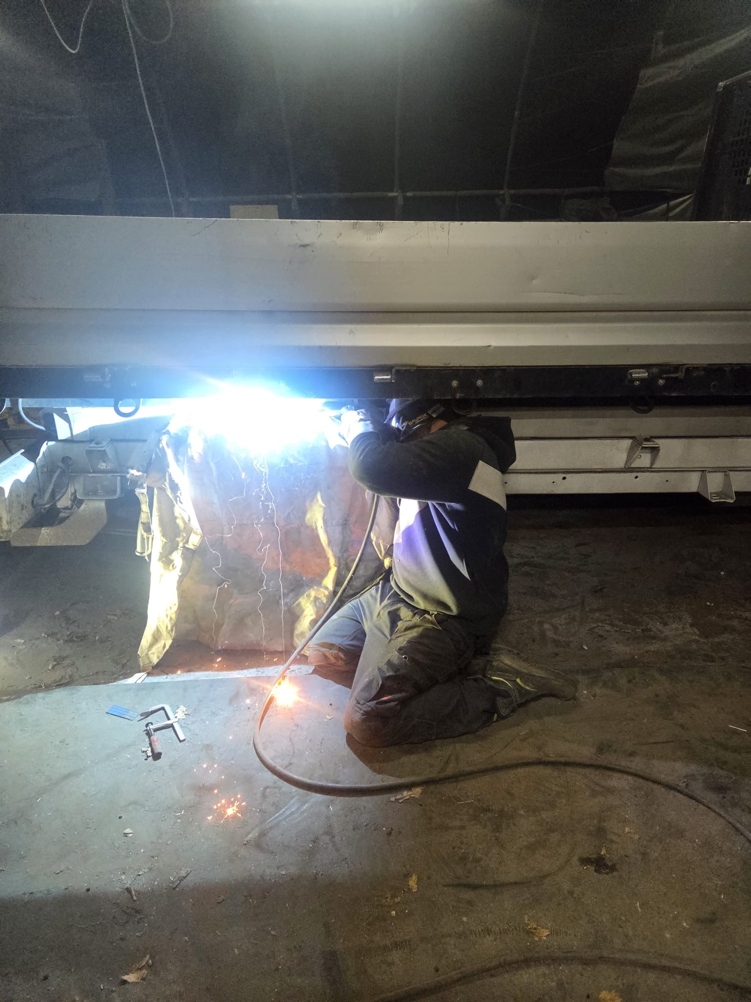 Spawacz w kombinezonie spawalniczym, klęczący pod konstrukcją, wykonuje spawanie. Widoczne iskry i intensywne światło łuku spawalniczego. Na podłodze leżą narzędzia.