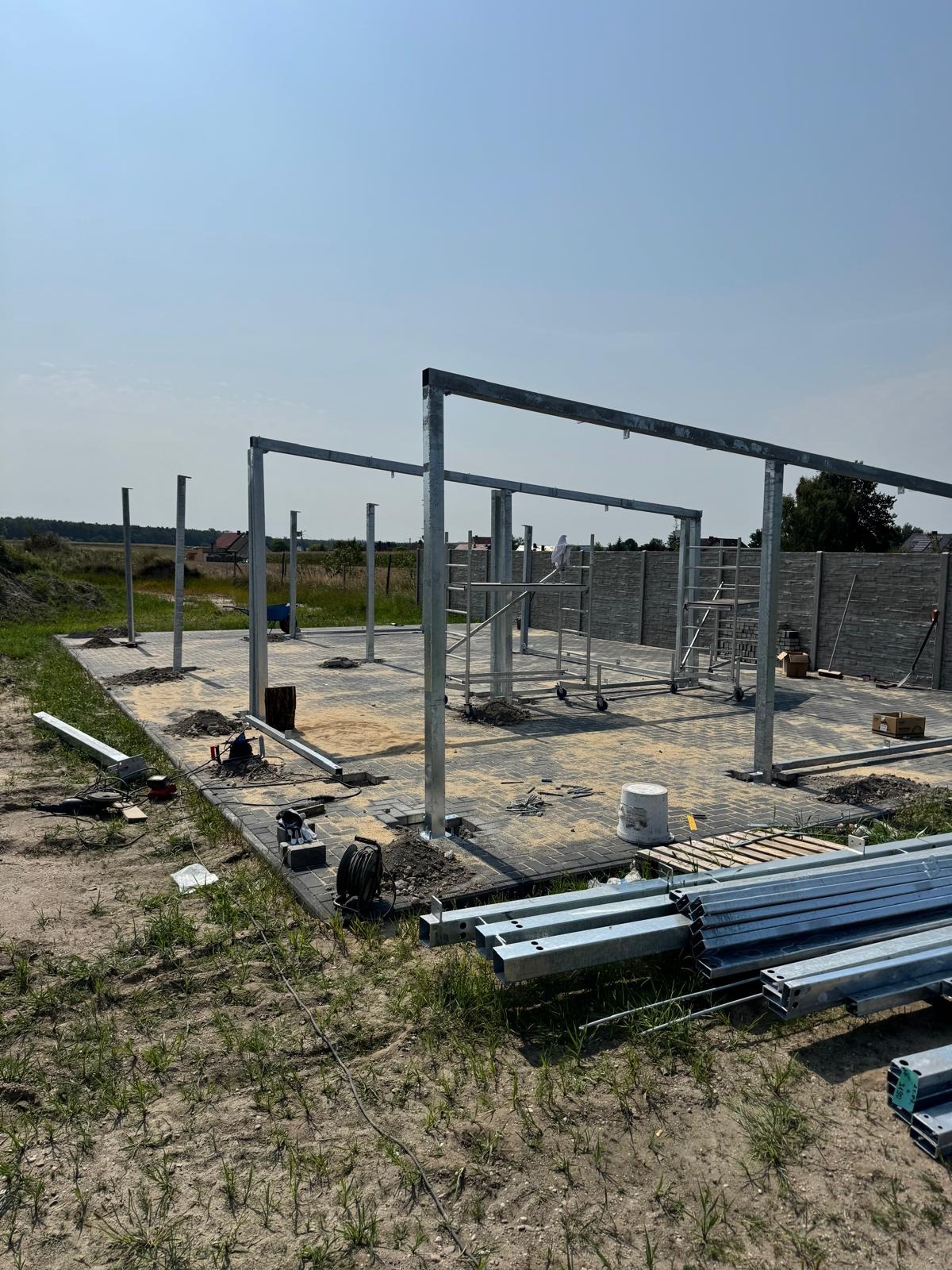 Montaż szkieletu hali z metalowych profili na fundamencie z kostki brukowej. Widoczne narzędzia i rusztowania. Słoneczny dzień, błękitne niebo.