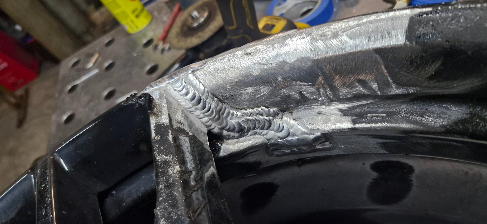 Detal spoiny na czarnej metalowej konstrukcji, widoczne ślady obróbki i narzędzia w tle. Precyzyjne łączenie elementów, surowy wygląd metalu.