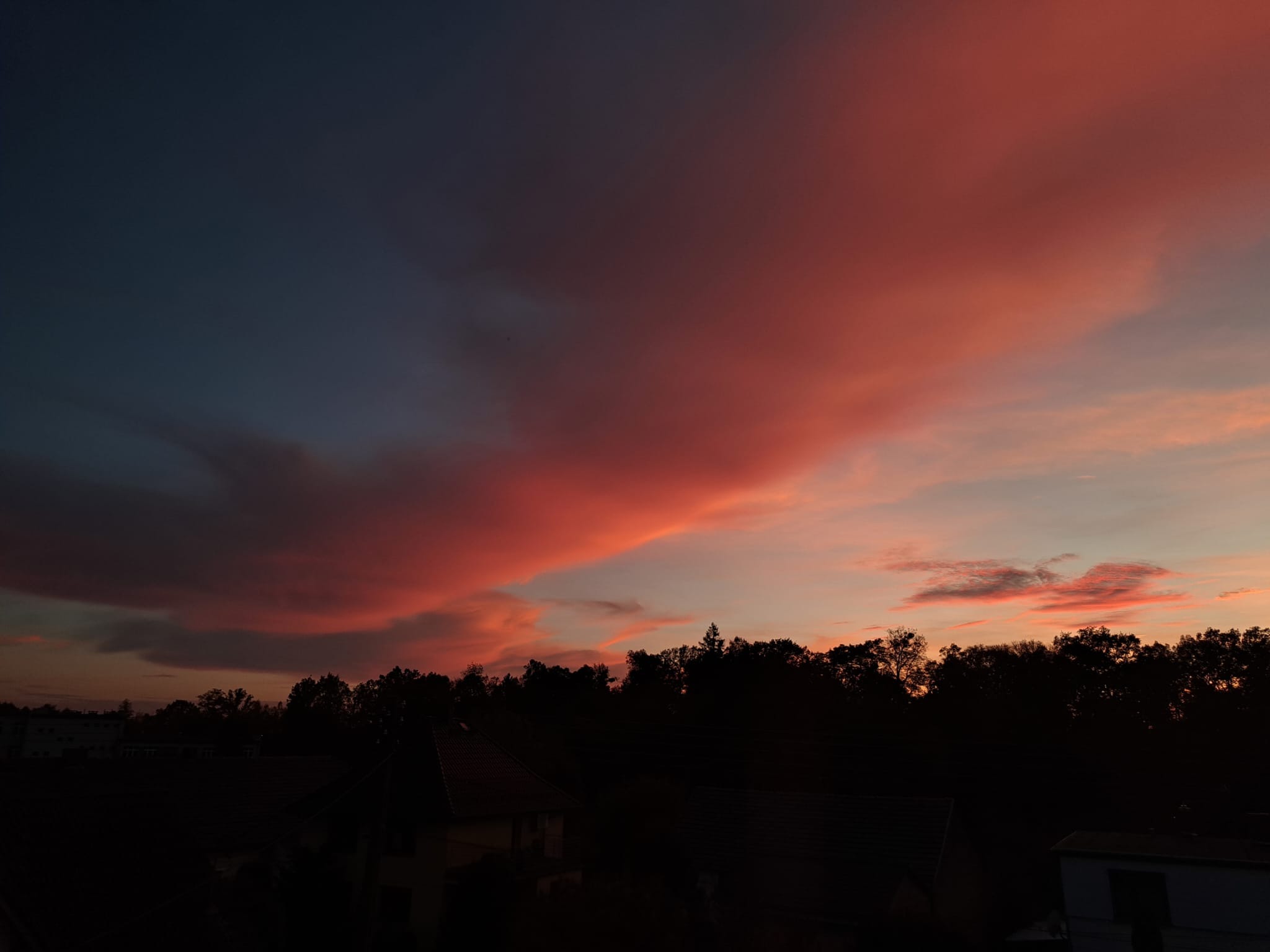 Wieczorny krajobraz z ciemnymi sylwetkami drzew i budynków na tle intensywnie różowego, zachodzącego słońca, tworzącego malownicze, rozmyte obłoki na niebie.