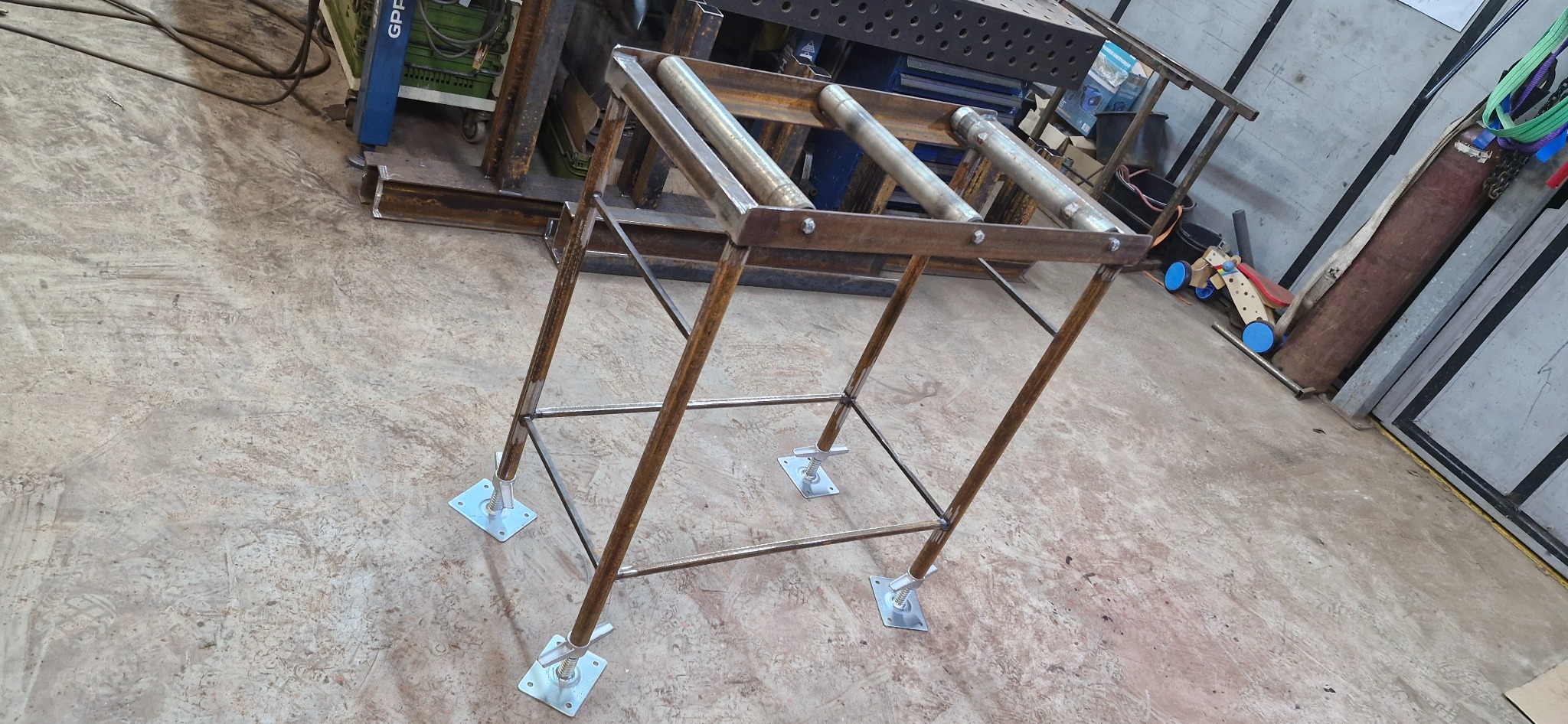 Metalowy stojak rolkowy z regulowanymi nóżkami, spawana konstrukcja, widoczne ślady obróbki, surowy wygląd, warsztatowe otoczenie.