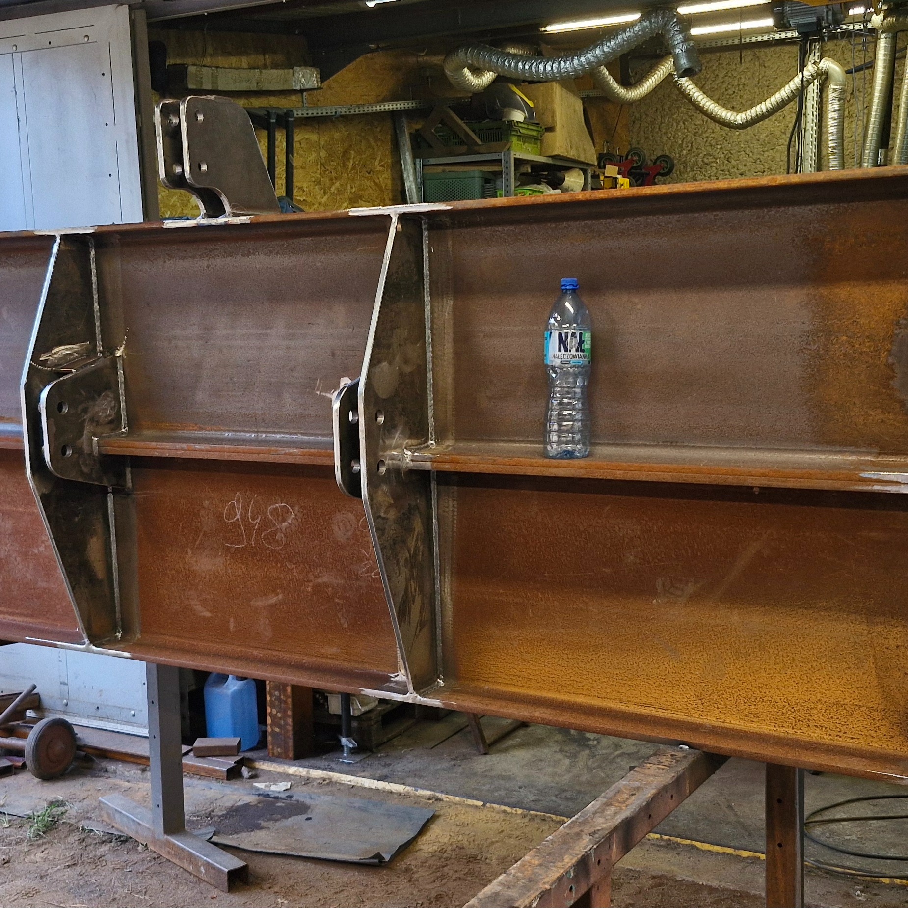 Fragment konstrukcji stalowej z widocznymi spawami i elementami mocującymi. Na konstrukcji stoi butelka wody. Widoczne ślady rdzy i niedoskonałości powierzchni.