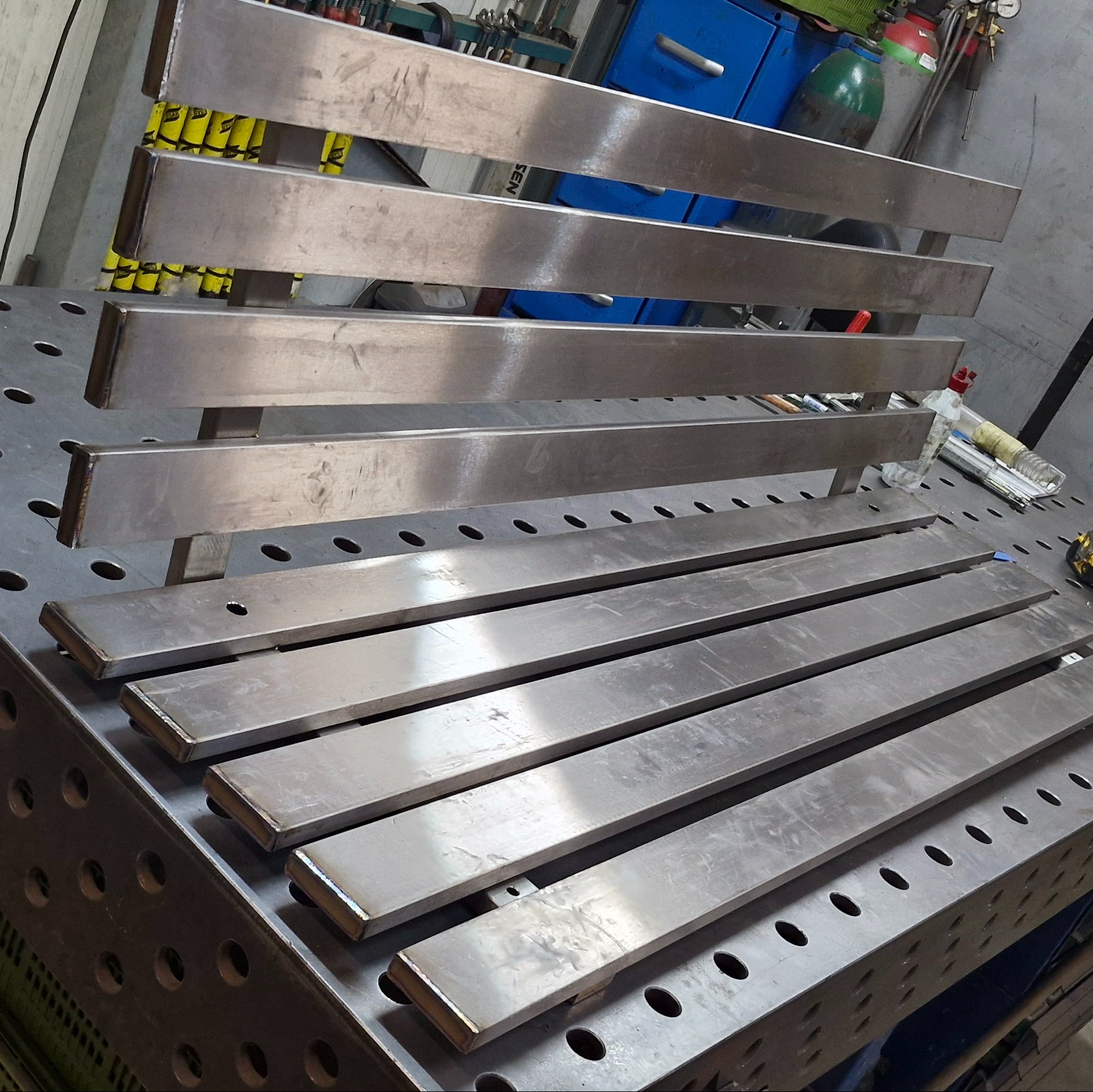 Metalowe elementy konstrukcyjne, prawdopodobnie nierdzewne, ułożone na perforowanym stole warsztatowym. Widoczne ślady obróbki i spawania. W tle narzędzia i szafka.