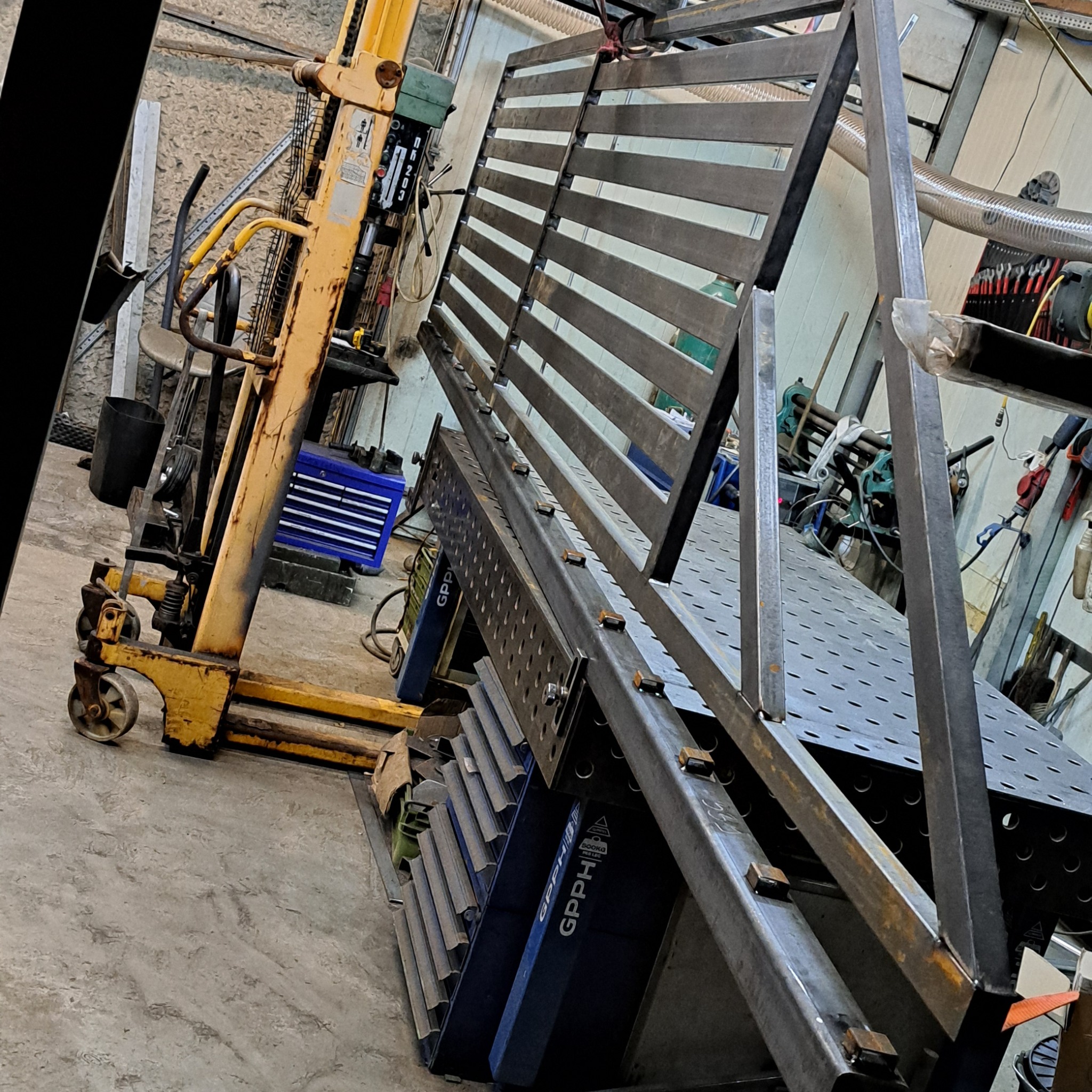 Metalowa konstrukcja z poziomymi szczeblami, podparta na niebieskich stojakach, w warsztacie. W tle widać wózek widłowy i narzędzia.