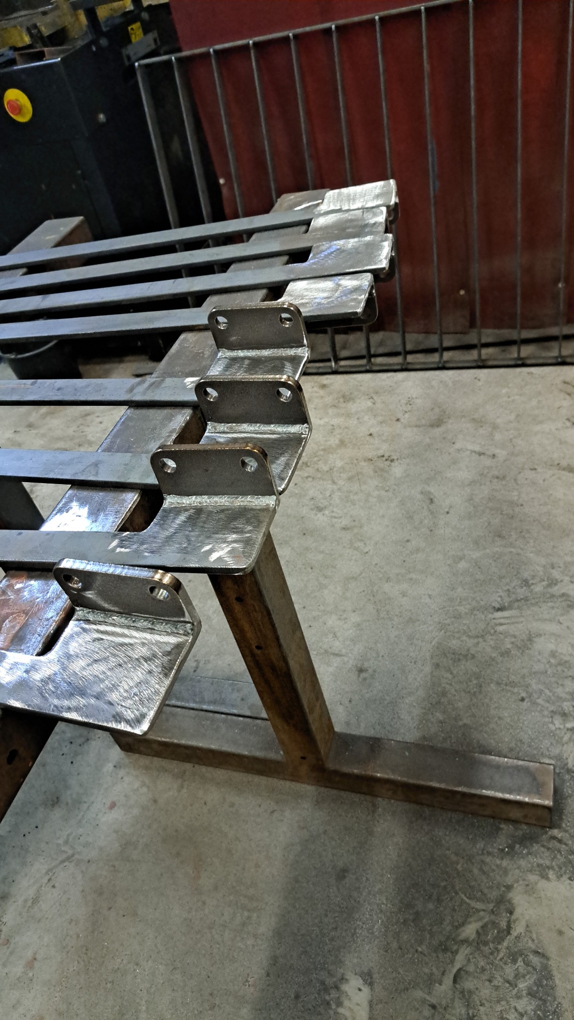 Metalowe elementy konstrukcyjne z otworami, ułożone na metalowym stojaku. Widoczne ślady obróbki i spawania. Tło stanowią metalowe pręty i maszyna.