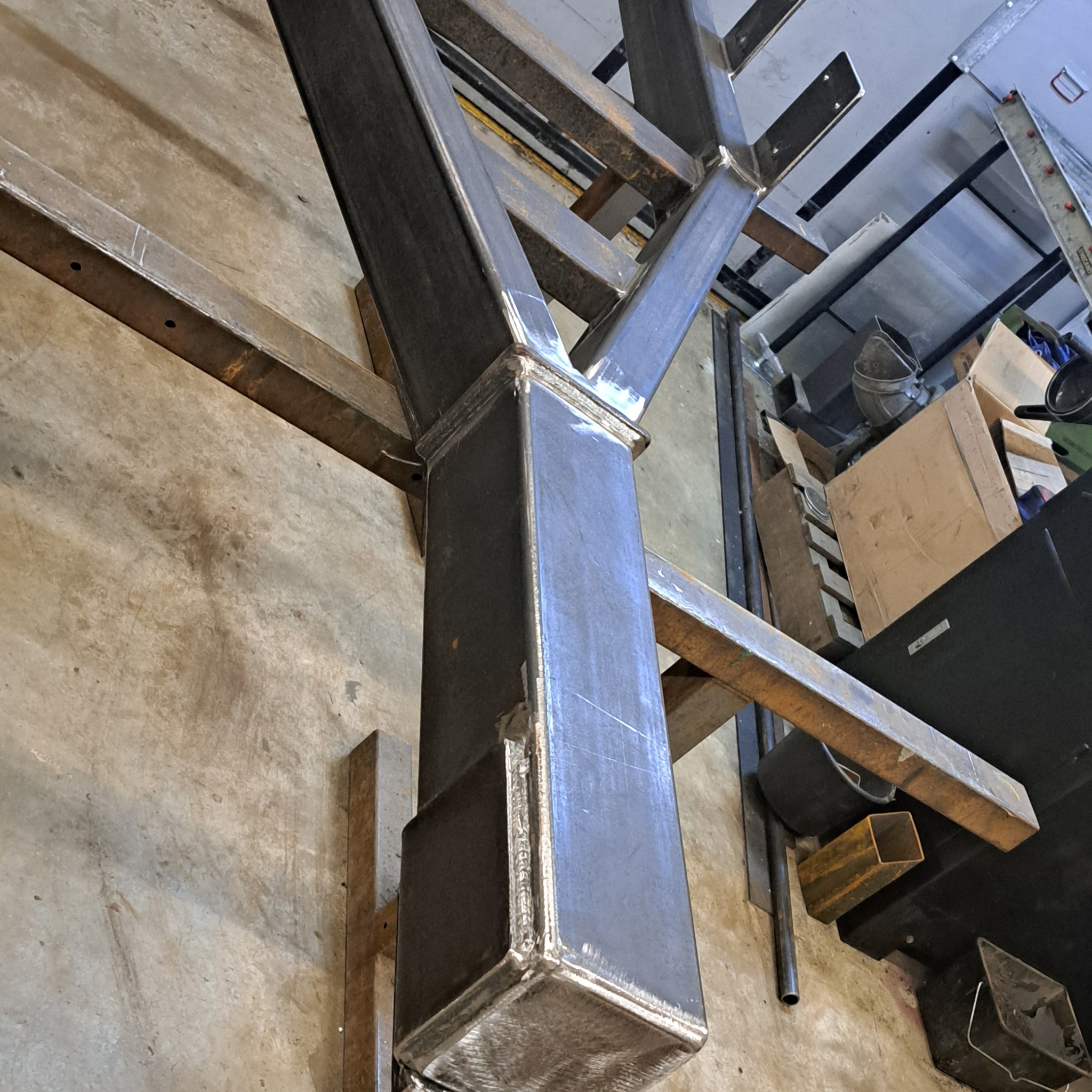 Konstrukcja metalowa z profili kwadratowych, spawana, surowa, w warsztacie. Widoczne spawy i elementy mocujące. Ujęcie z dołu, podkreślające solidność konstrukcji.