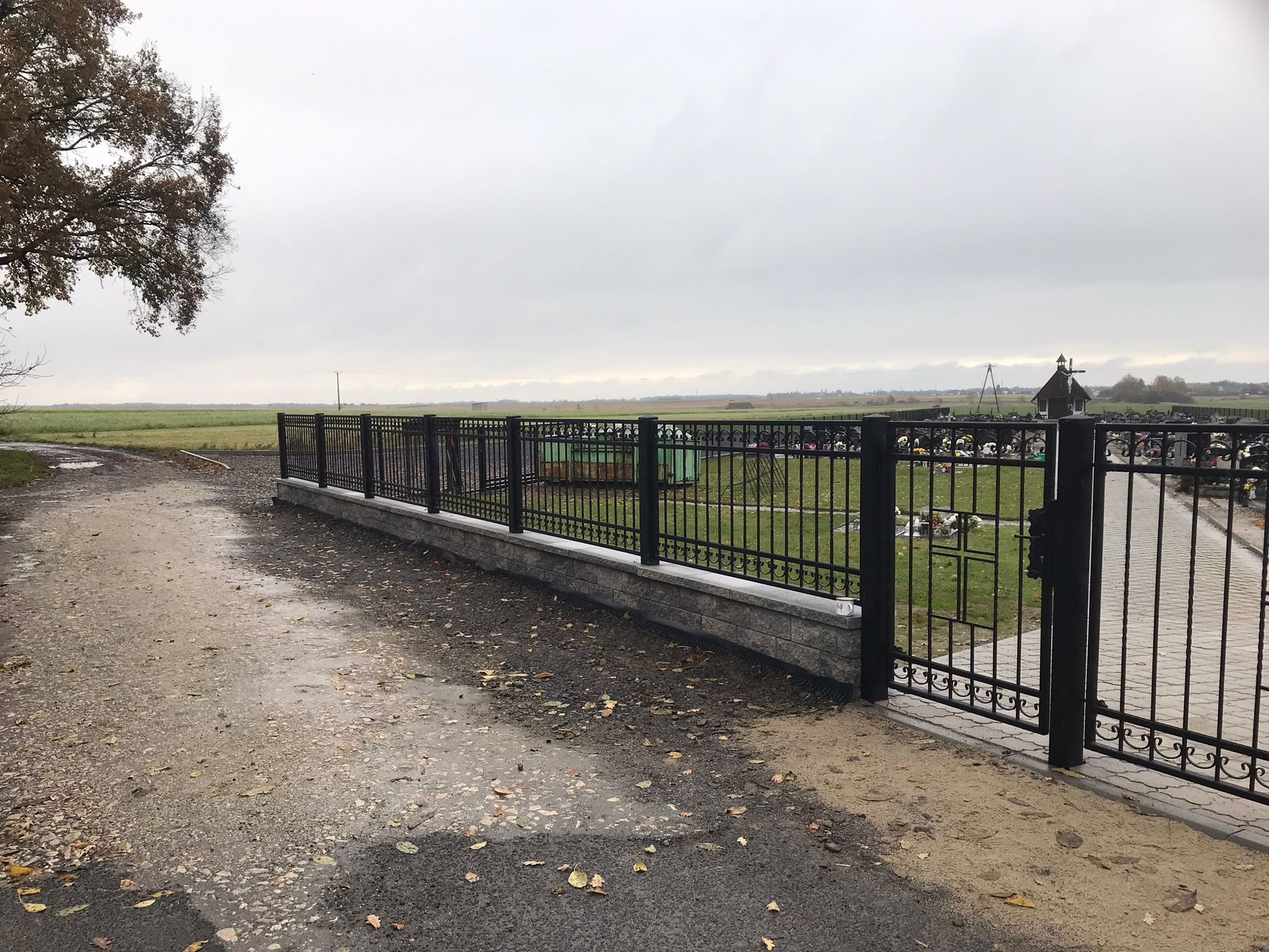 Czarne, metalowe ogrodzenie z bramą, oddzielające cmentarz od polnej drogi. Widoczne nagrobki i kapliczka w tle. Pochmurne niebo.