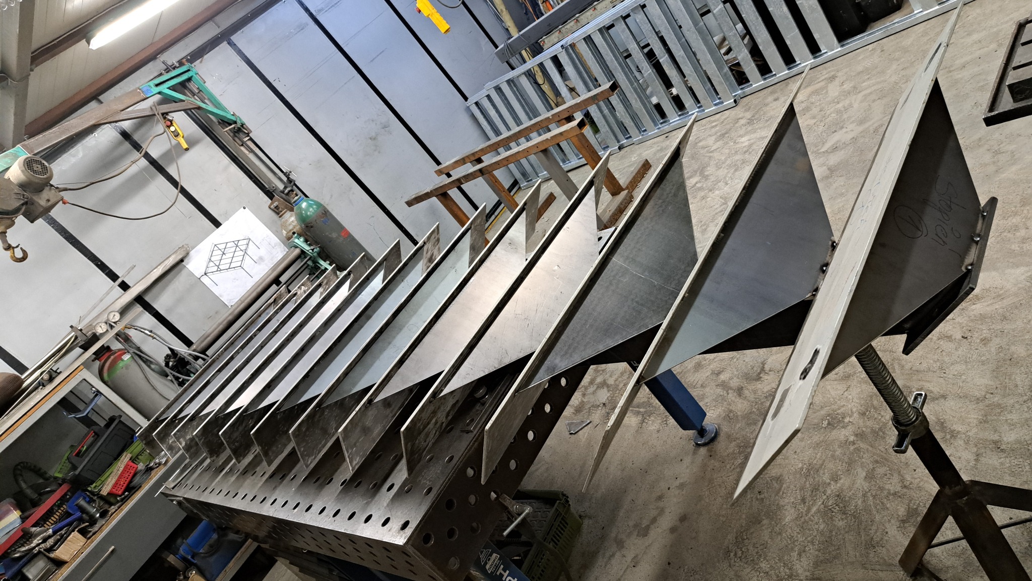 Metalowe elementy konstrukcyjne, prawdopodobnie do hali, ustawione rzędem na perforowanym stole spawalniczym. Widoczne spawane łączenia i podpory. Warsztat ślusarski.