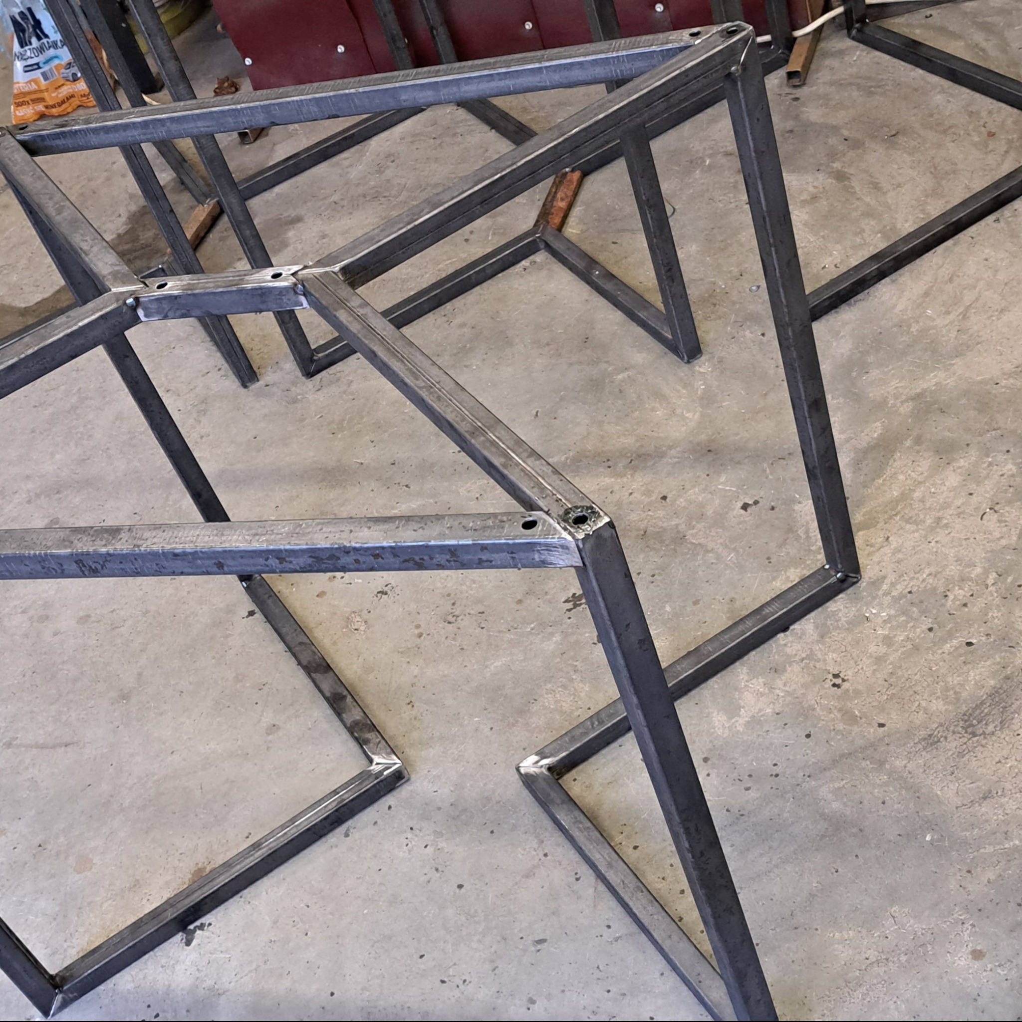 Metalowe, spawane konstrukcje w kształcie sześcianów, przygotowane do dalszej obróbki. Widoczne spawy i otwory montażowe. Surowy, warsztatowy charakter.