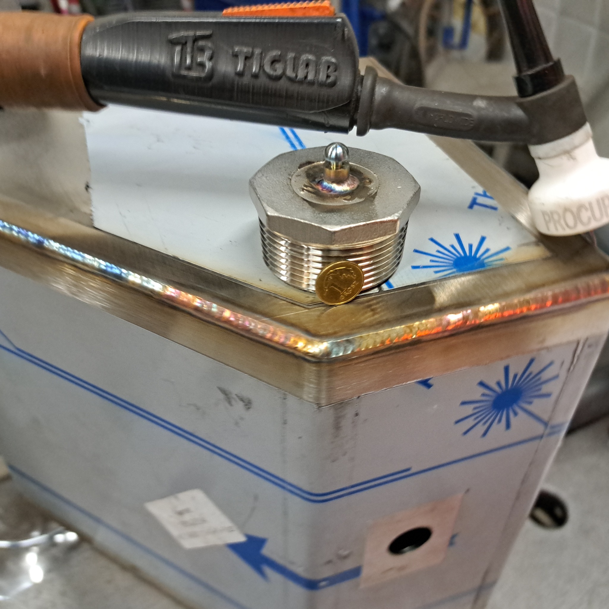 Detal spoiny TIG na zbiorniku ze stali nierdzewnej z monetą dla skali, widoczny uchwyt palnika TIGLAB. Precyzyjne łączenie blach, widoczne efekty termiczne.