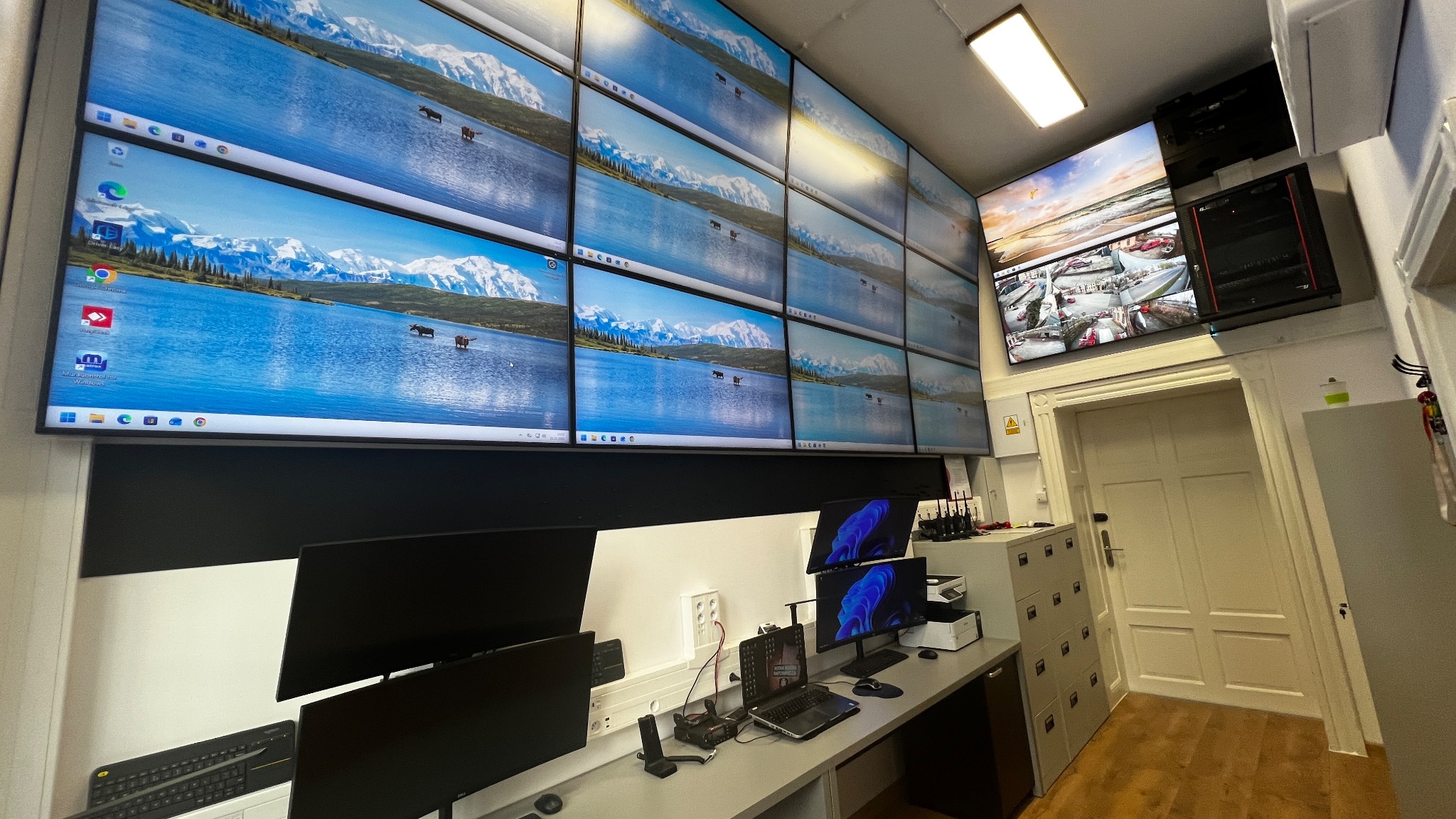 Sala monitoringu z wieloma ekranami wyświetlającymi obraz z kamer i pulpity komputerów. Widoczne kable, gniazda i sprzęt IT. Skonfigurowany system monitoringu.