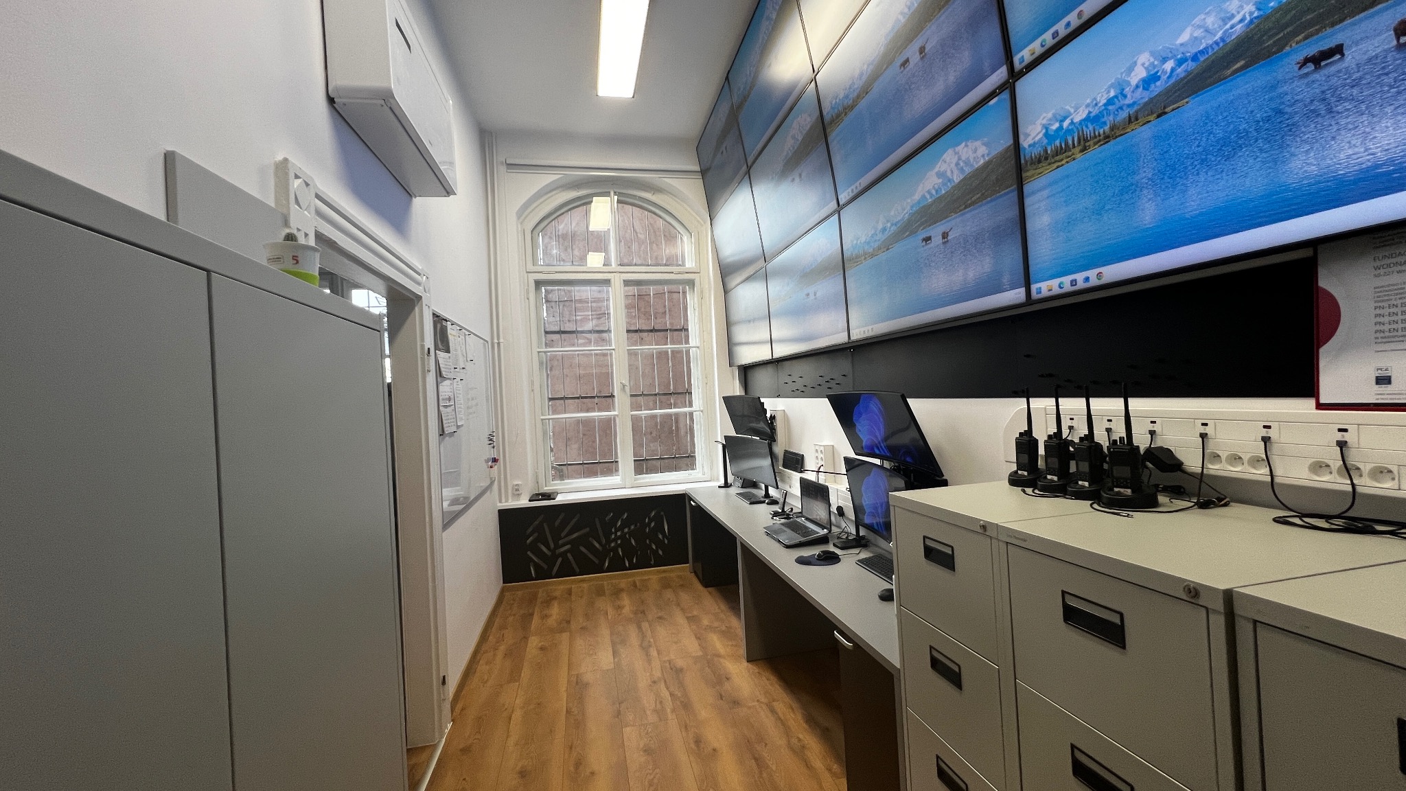 Centrum monitoringu z wieloma ekranami wyświetlającymi krajobraz, biurkiem z komputerami i radiotelefonami. Nowoczesne wyposażenie, jasne oświetlenie, parkiet.