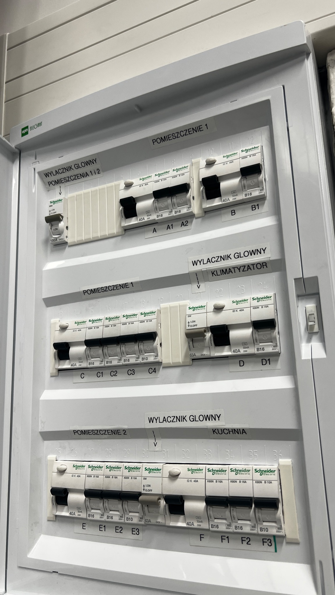 Rozdzielnica elektryczna z bezpiecznikami Schneider, opisana dla pomieszczeń: 1, 2, kuchni i klimatyzatora. Widoczne wyłączniki główne i poszczególne obwody.