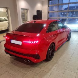 My Clean. Car Premium Detailing - Czerwone Audi RS3 po oklejeniu folią, widoczne czarne felgi i przyciemnione szyby. Auto stoi w jasnym pomieszczeniu z przeszkloną ścianą.