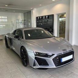 My Clean. Car Premium Detailing - Srebrne Audi R8 na tle jasnego salonu z przeszklonymi ścianami i czarnymi półkami. Sportowy samochód z czarnymi felgami prezentuje się elegancko.